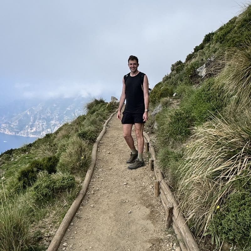 Brett mid-hike on a narrow cliffside path, fog hanging over the sea far below