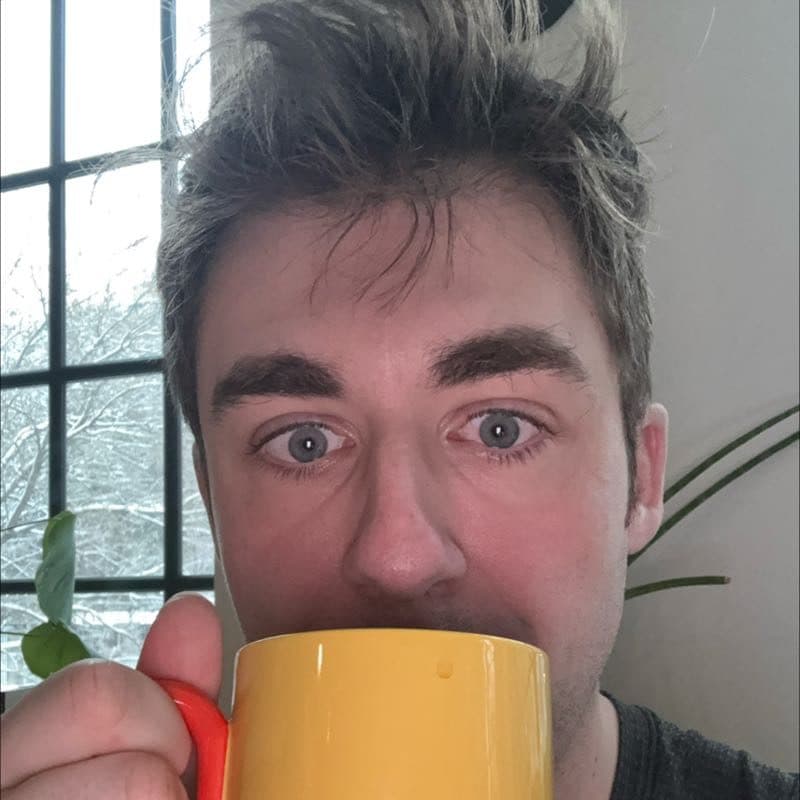 Brett peering over the rim of a big yellow mug, snow out the window behind him