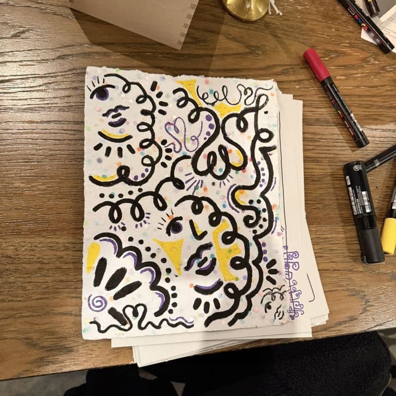 A sheet of handmade paper covered edge to edge in looping marker doodles, paint pens scattered around